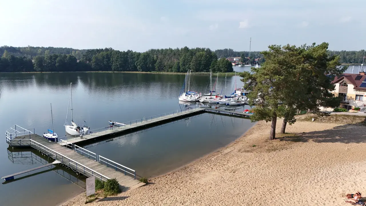 Wdzydze Kiszewskie plaża z pomostami i żaglówkami. Piasek, woda i zielone drzewa tworzą idylliczny krajobraz.