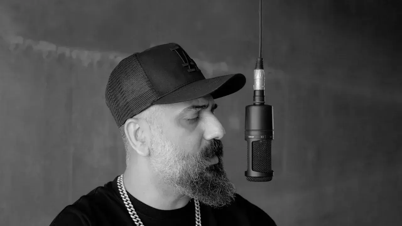 Artysta w czapce z daszkiem i łańcuchem na szyi nagrywa utwór rnb przy mikrofonie.