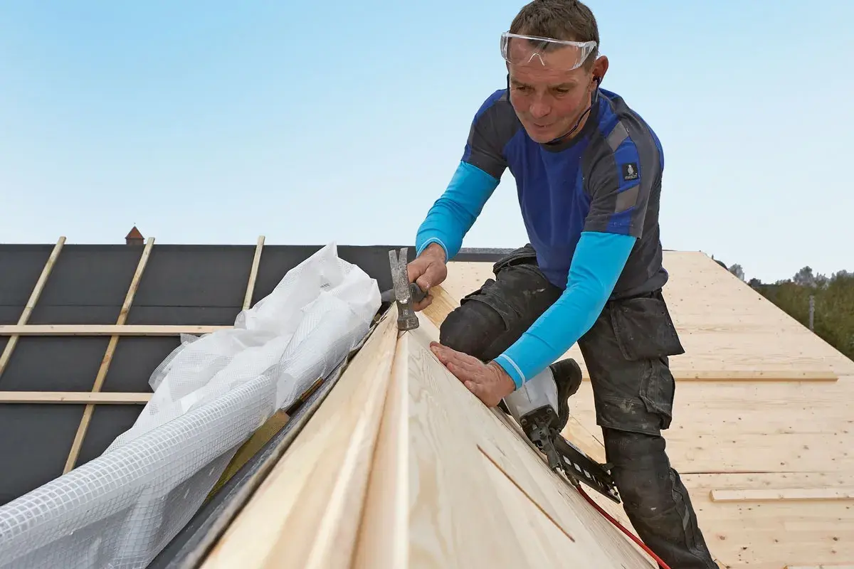 A roofer wearing safety glasses hammers a wooden plank onto a roof under construction.