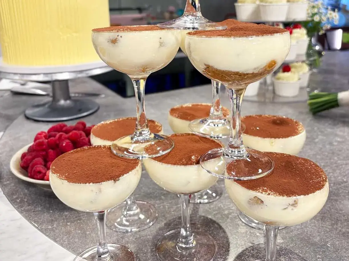Flera glas med krämig tiramisu, pudrad med kakao. Hallon och en gul tårta i bakgrunden.