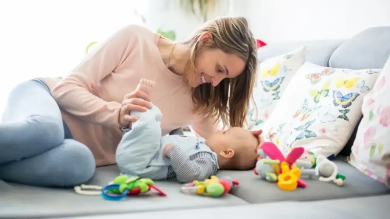 Mamá sonríe a su bebe de 2 meses mientras juegan en el sofá entre coloridos juguetes.