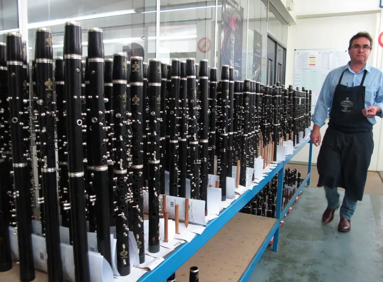 Un homme en tablier inspecte un buffet de clarinettes, prêt à être expédié. Ces instruments de musique, des modèles "crampon mantes", sont alignés sur des étagères.
