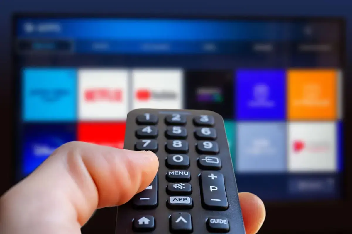 Hand håller fjärrkontroll framför en TV-skärm med app-ikoner. Kanske dags att välja en film eller en dokumentär om ze rappare.