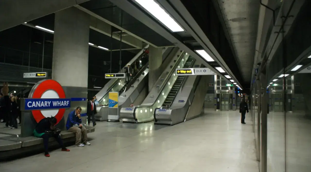 Stacja Canary Wharf w **metro londyn** z charakterystycznym znakiem, ruchomymi schodami i ludźmi czekającymi na pociąg.