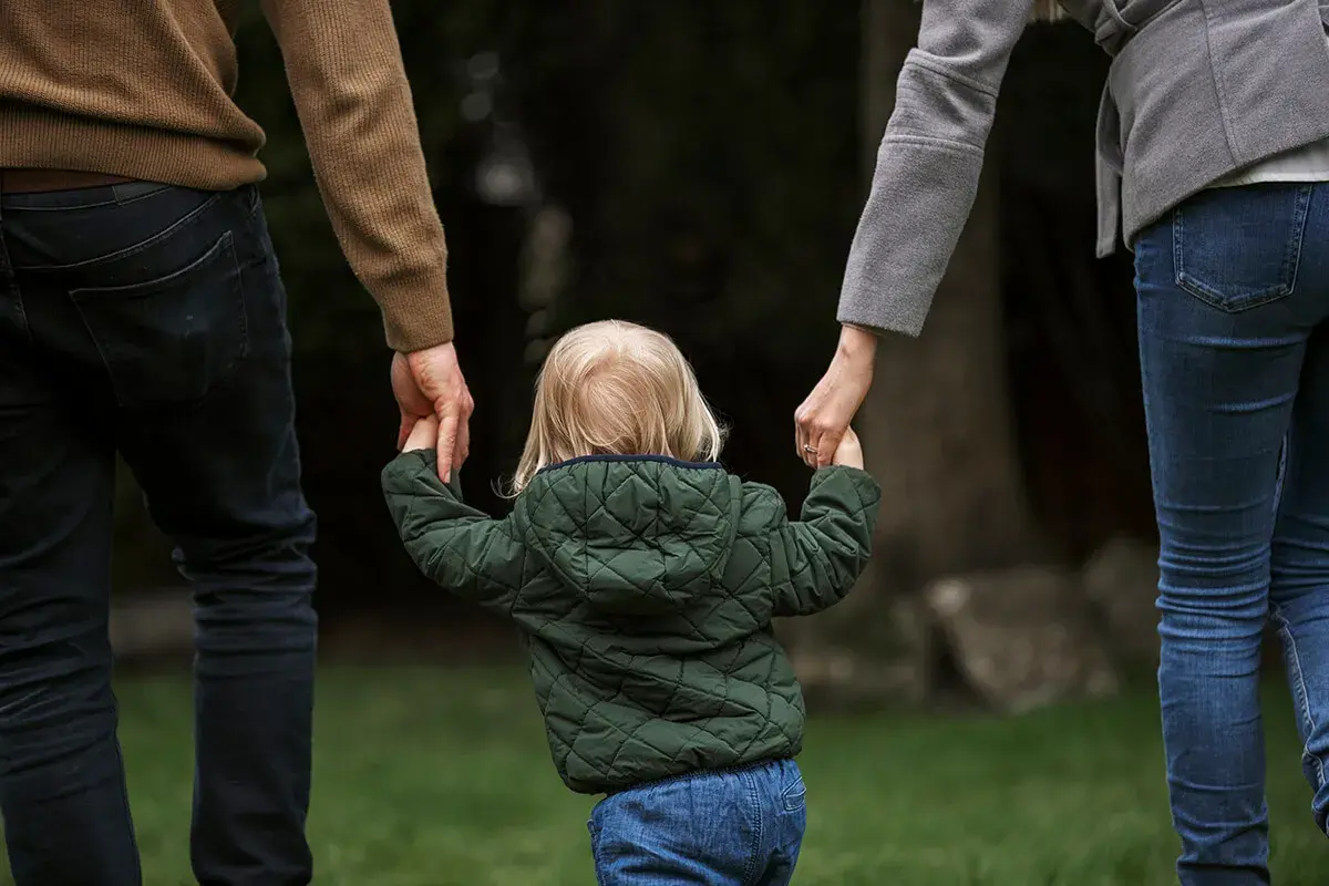 Dziecko z blond włosami, w zielonej kurtce, trzyma za ręce rodziców. To obraz przysposobienia niepełne, pełnego miłości i nadziei.