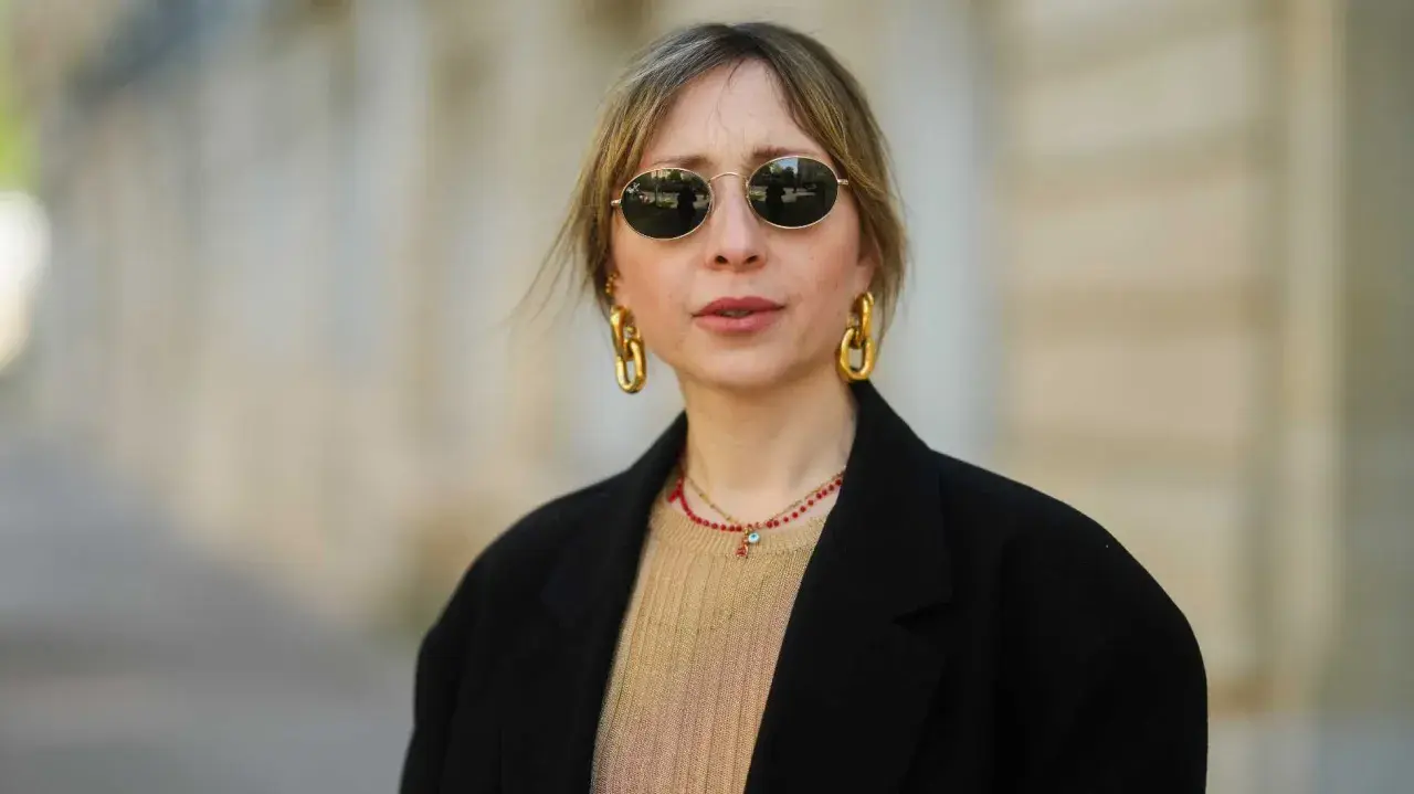 Frau mit stylischer curtain bangs frisur, runden Sonnenbrillen und auffälligem Goldschmuck.