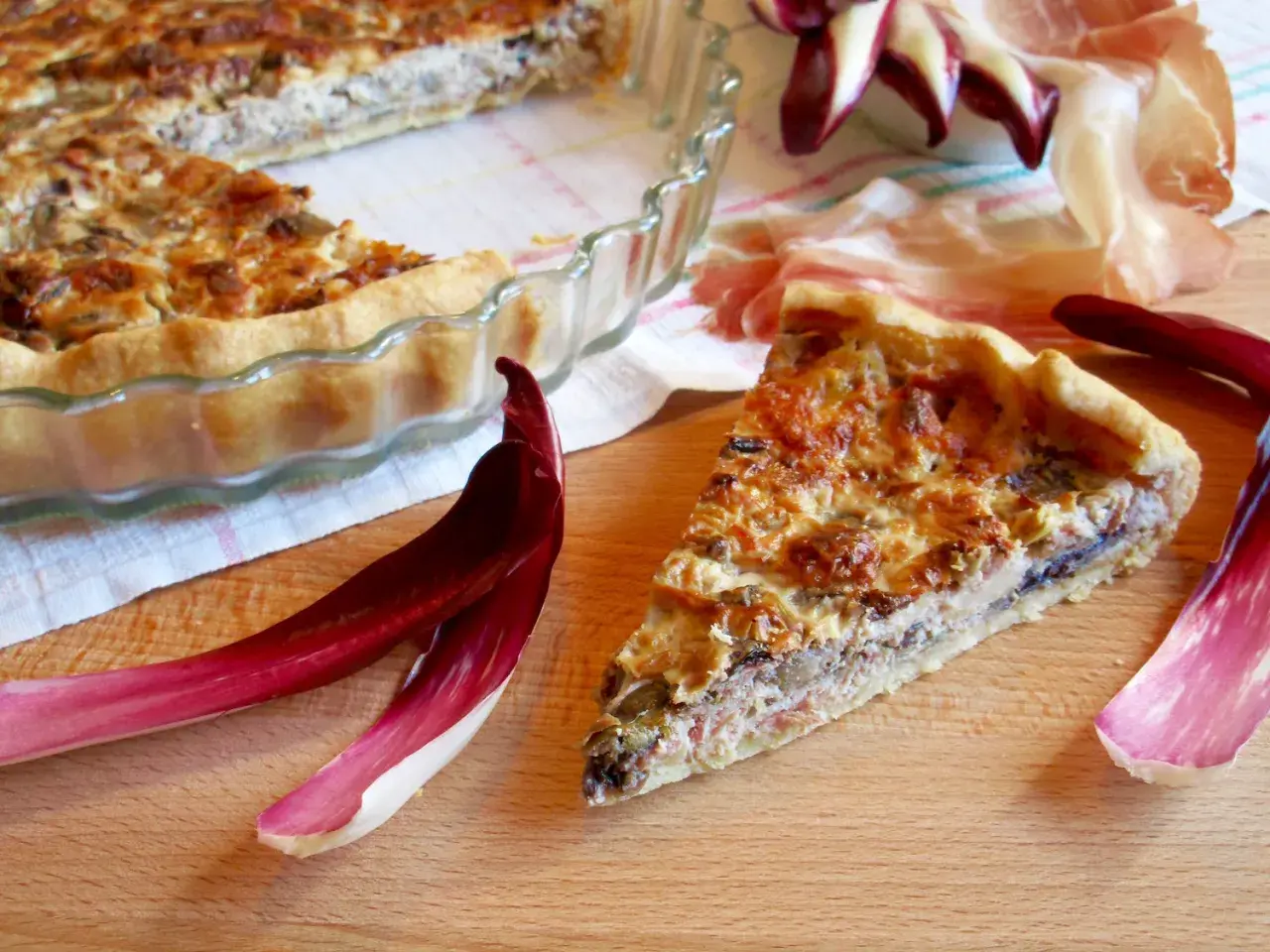Una fetta di torta salata con taleggio, ricca e dorata, accanto a foglie di radicchio e prosciutto crudo.