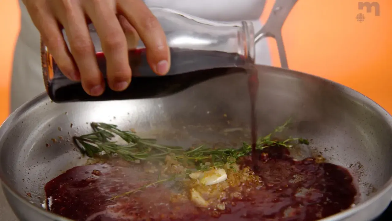 Une main verse du jus de viande dans une poêle avec de l'ail et des herbes.
