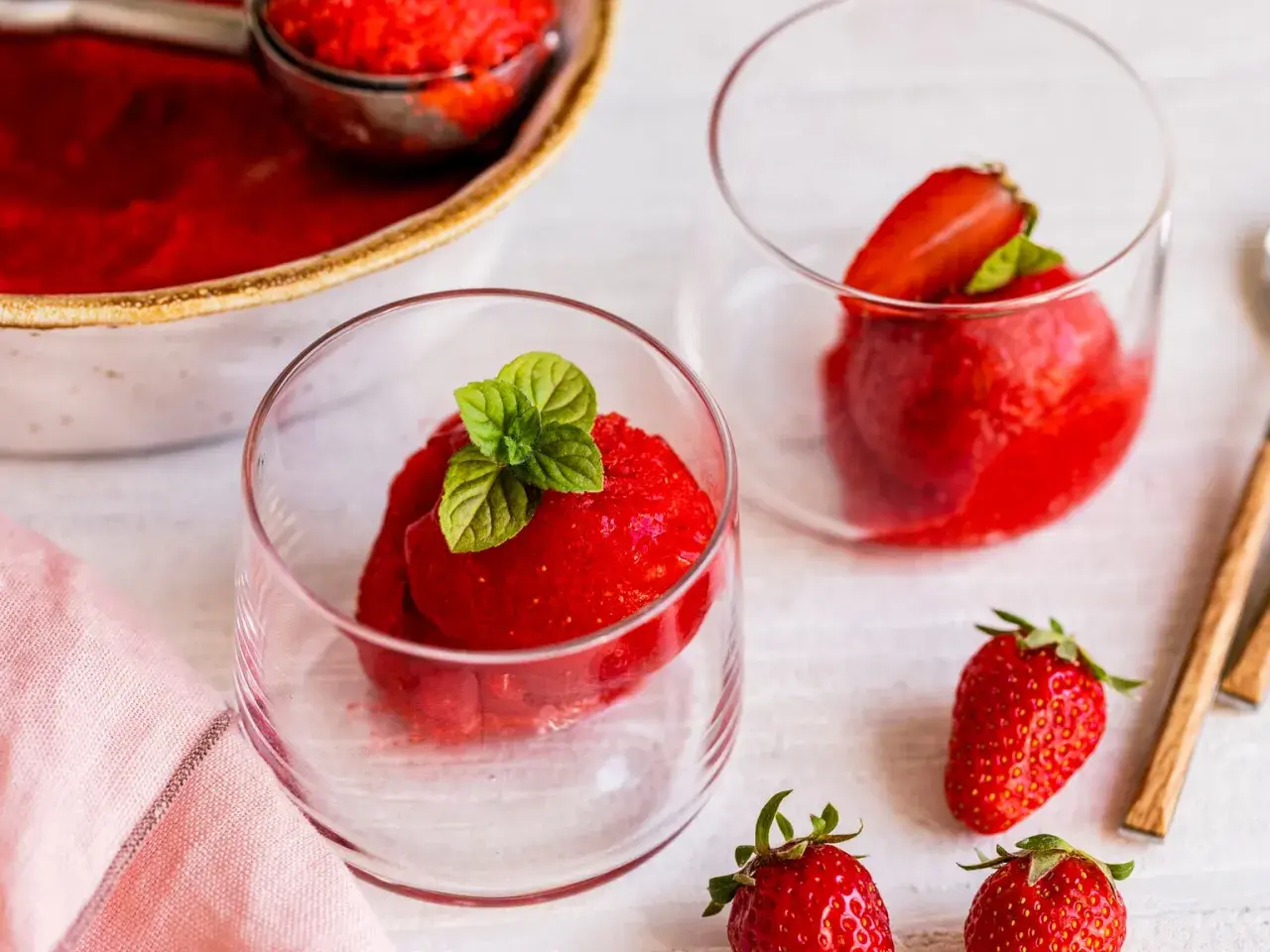 Erfrischendes Erdbeer Sorbet in Gläsern, garniert mit Minze. Daneben frische Erdbeeren und eine Schüssel mit dem leuchtend roten Sorbet.
