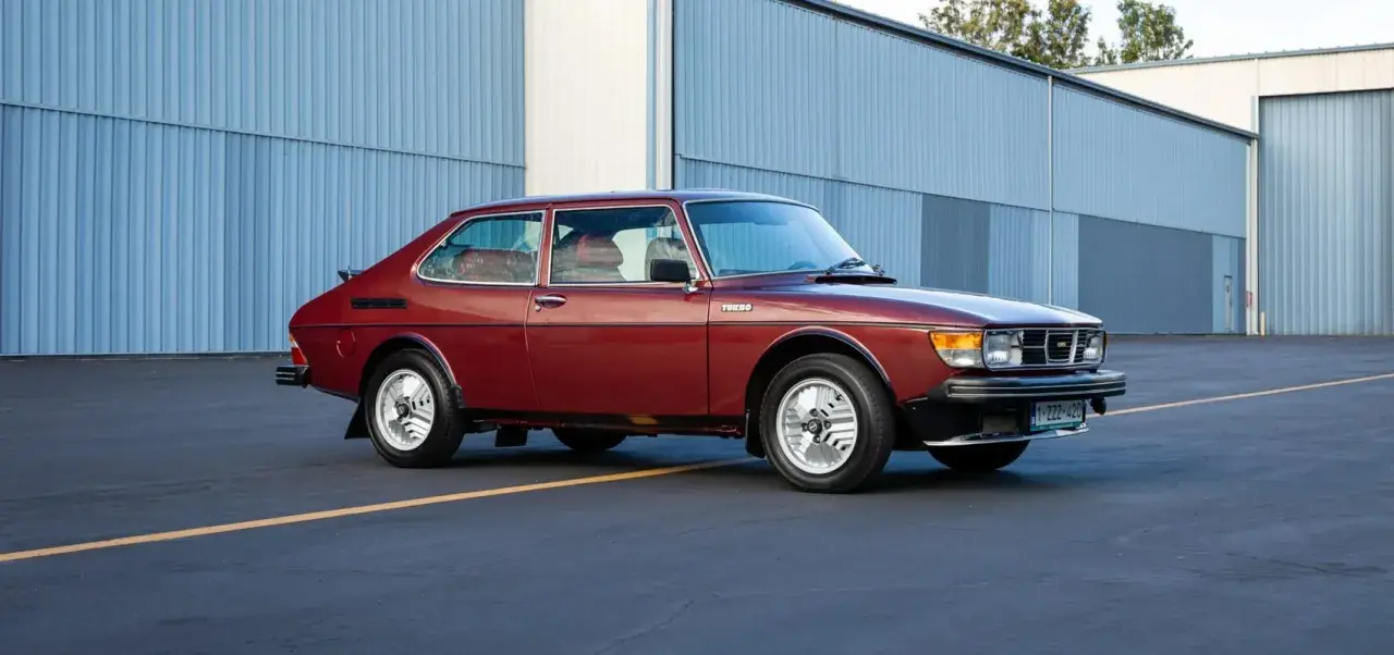 En röd Saab 99 Turbo står parkerad framför en blå hangar.