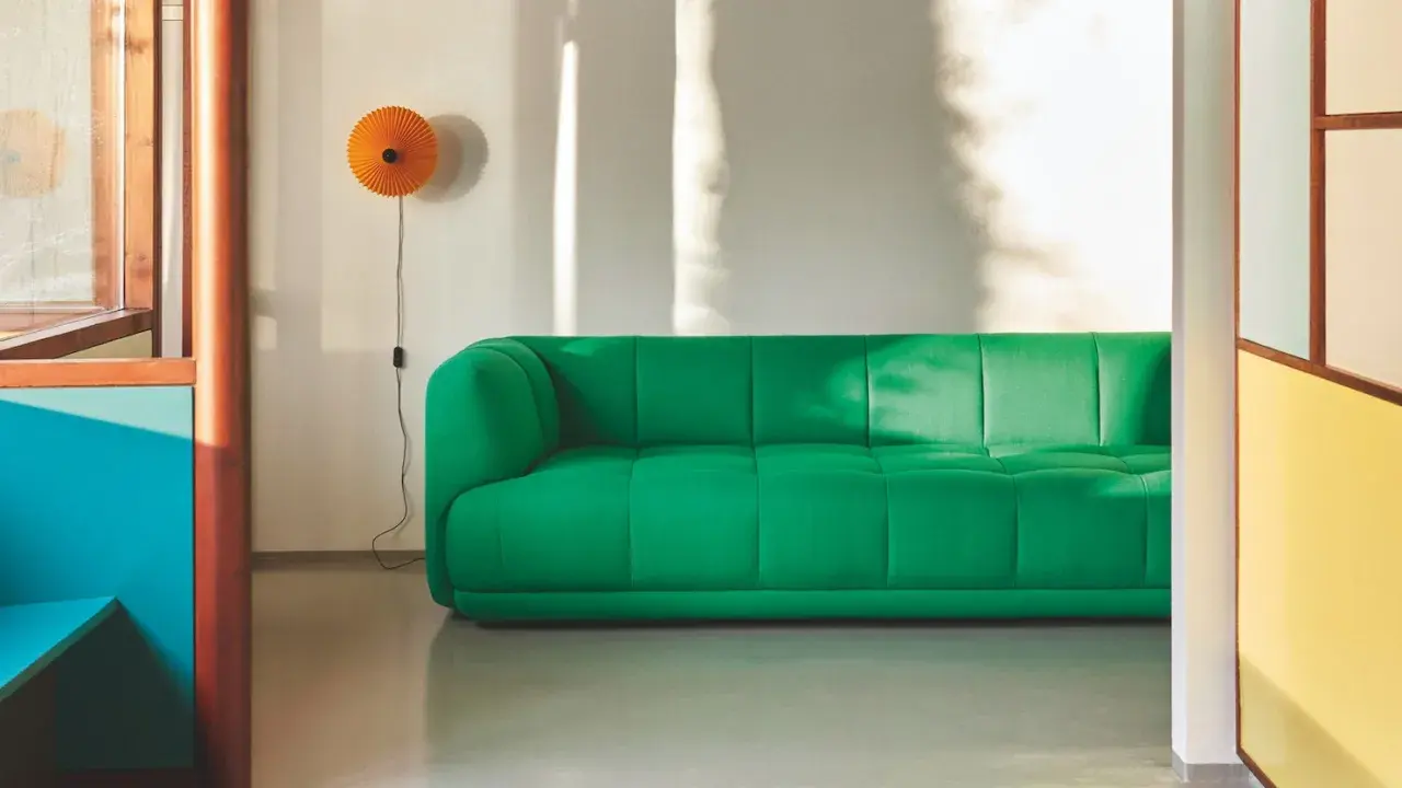 Ein leuchtend grünes Sofa steht vor einer weißen Wand, an der eine orangefarbene Lampe hängt. Sonnenlicht wirft Schatten. Mette hay.