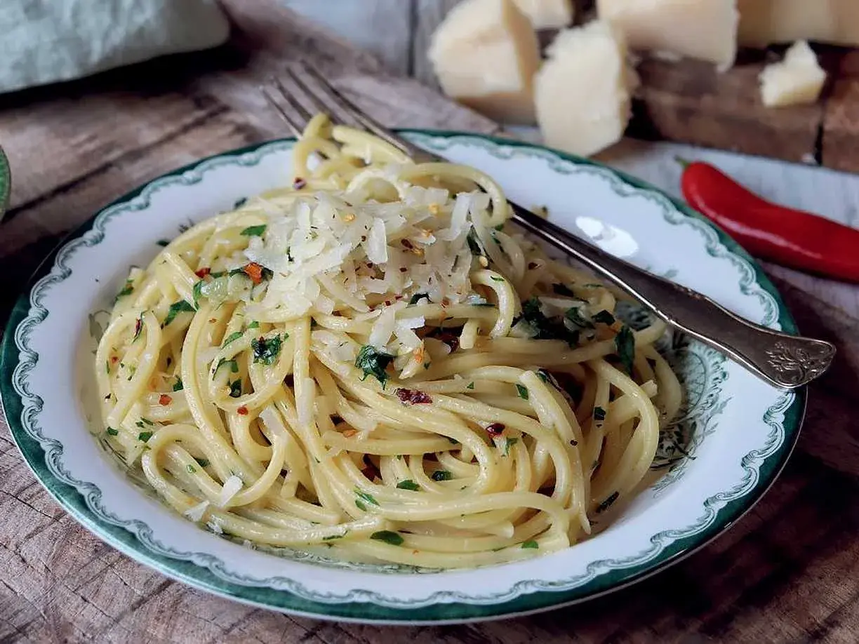 En tallrik med spaghetti aglio e olio, toppad med riven parmesan och persilja. En röd chili och ostbitar syns i bakgrunden.