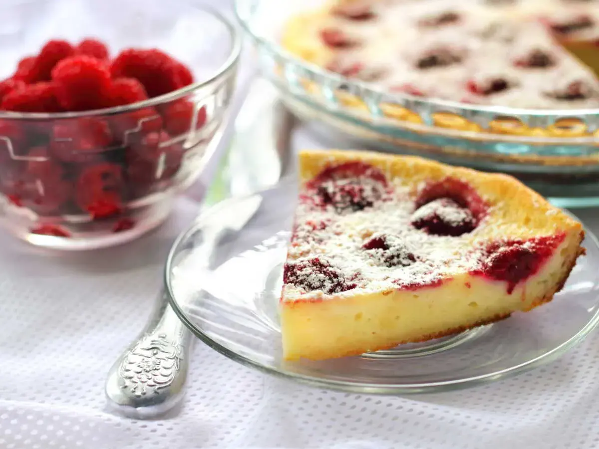 Une part de clafoutis framboise, saupoudrée de sucre glace, à côté d'un bol de framboises fraîches.