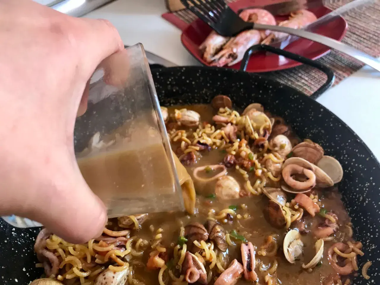 Vertiendo caldo en una paellera con fideos, calamares y almejas. Así se hace fideuá.