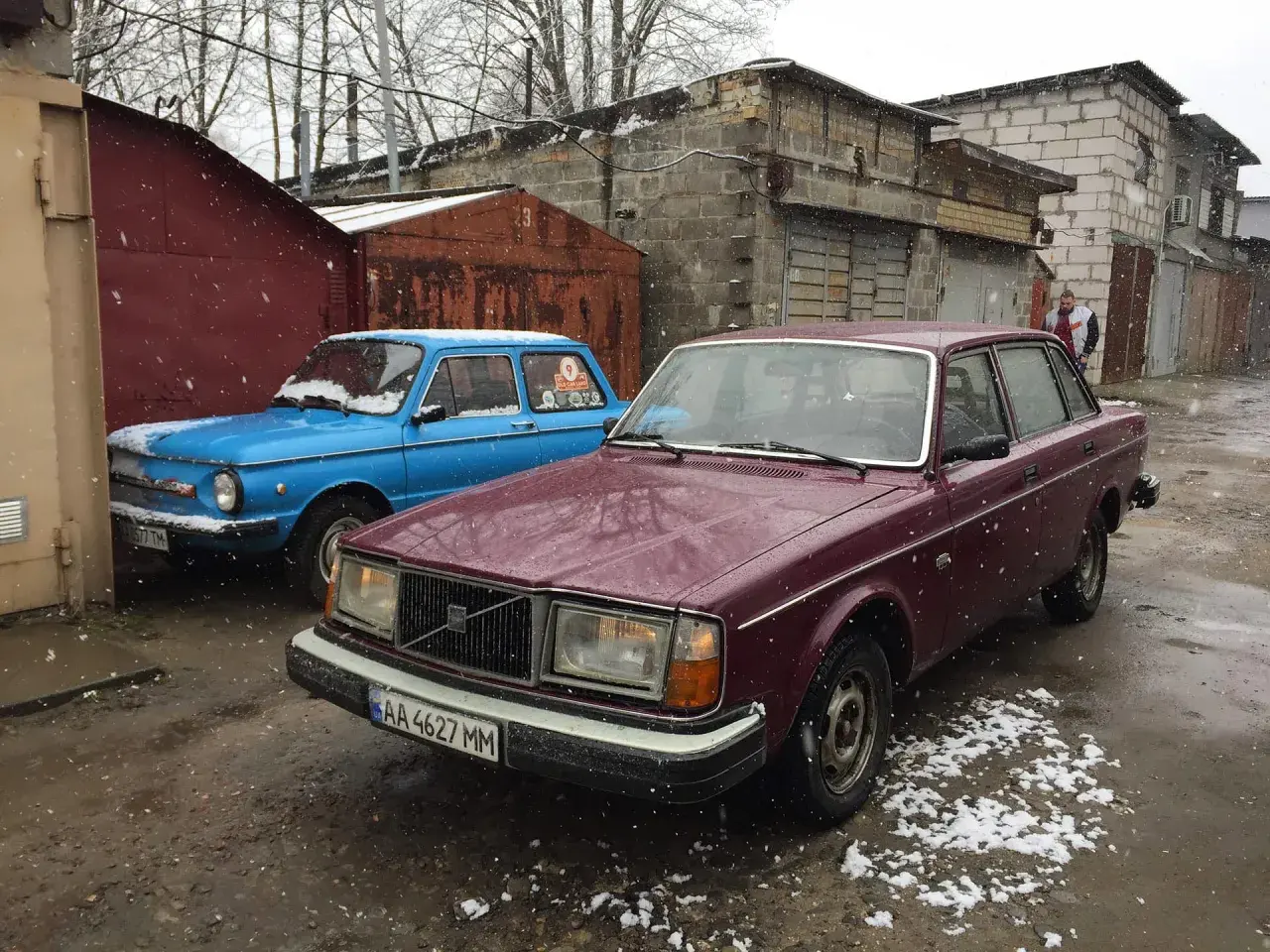 En vinröd Volvo 244 DLS står parkerad bredvid en blå bil i snöfall.
