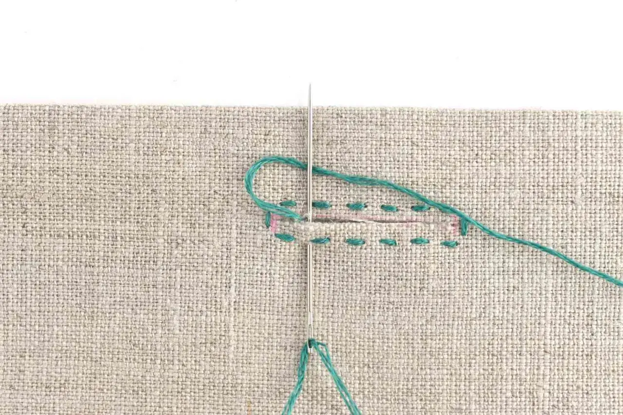 Close-up of a needle and teal thread being used to sew a buttonhole on beige fabric. Stitches are visible along the edges of the cut.