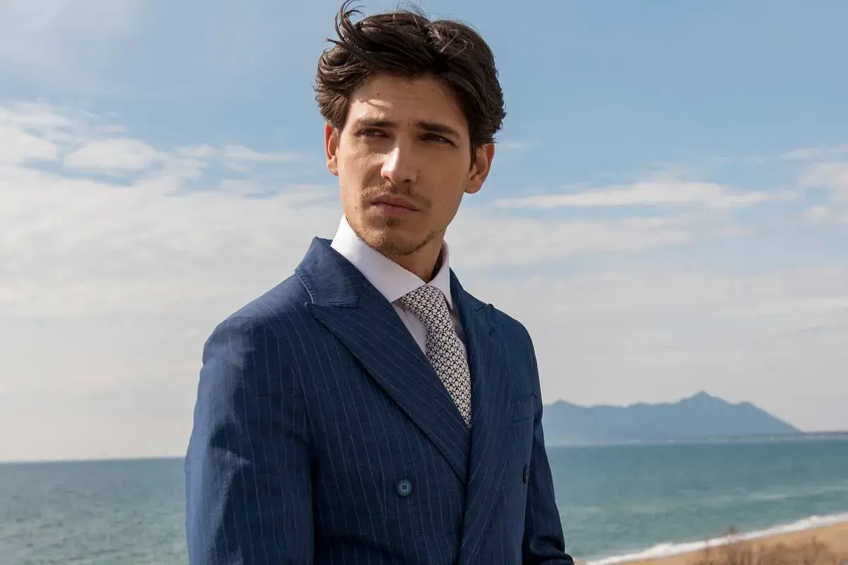 Uomo con abito blu gessato e camicia bianca, indossa una cravatta con un elegante nodo cravatta simmetrico. Sguardo pensieroso verso l'orizzonte marino.