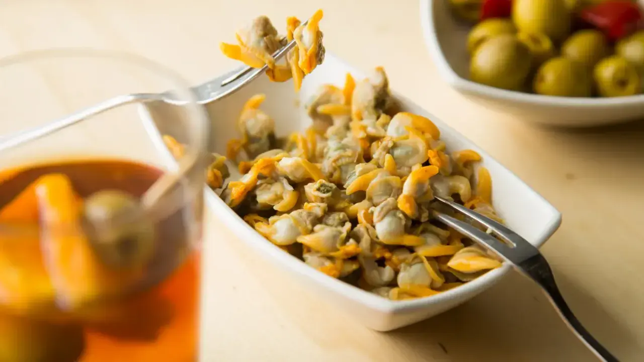 Un plato de berberechos, pequeños moluscos marinos, listos para disfrutar. Se ven deliciosos junto a un vaso de bebida y aceitunas.