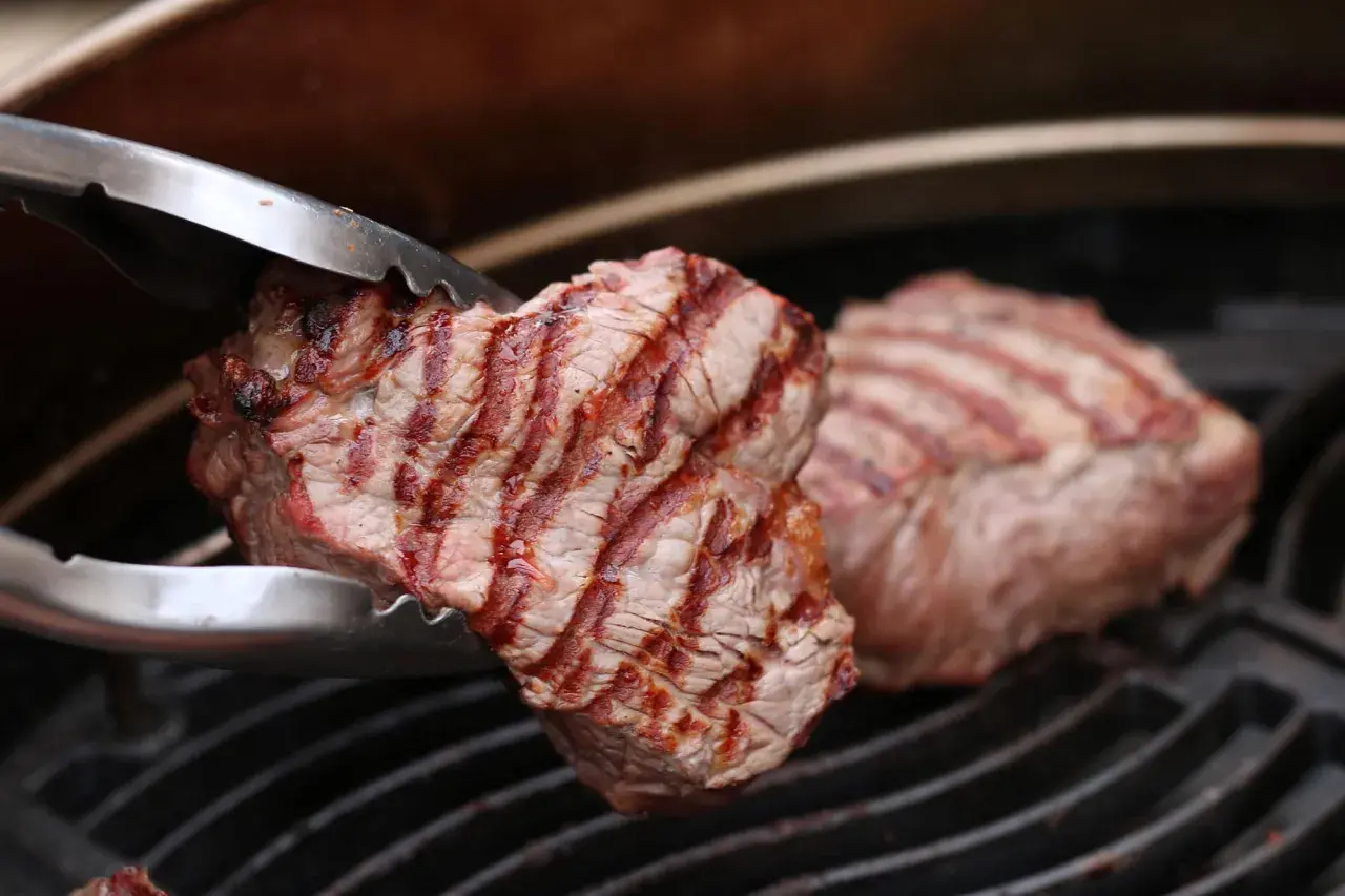 Zangen halten ein saftiges Stück Roastbeef über einem Grillrost. Das Fleisch hat schöne Grillstreifen und ist perfekt gegrillt.