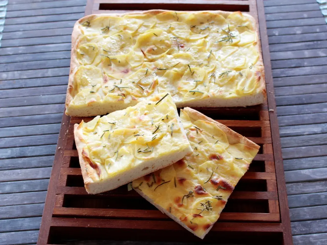 Focaccia con lievito istantaneo, ricoperta di patate sottili e rosmarino, tagliata a fette su un tagliere di legno.