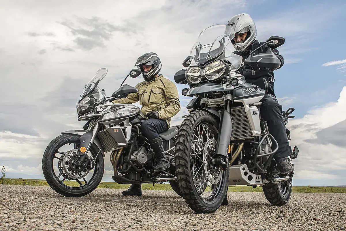 Dos motociclistas con cascos y equipo de aventura listos para explorar caminos y **trail asfalticas** bajo un cielo nublado.