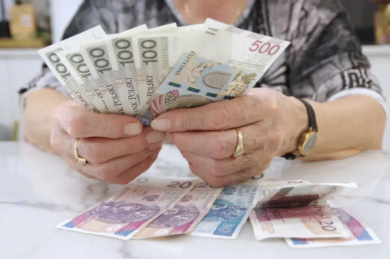 Dłonie z obrączkami trzymają banknoty 100 i 500 zł. Na stole leżą banknoty 20 zł. Czerwcowe emerytury już są!