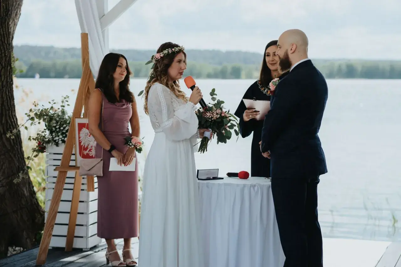 Panna młoda z mikrofonem i bukietem mówi podczas ceremonii. Obok stoi pan młody, urzędniczka i świadkowa. To ślub jednostronny nad jeziorem.