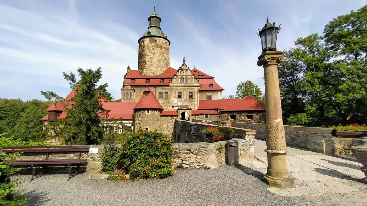 Zamek Czocha, gdzie jest? Malowniczy zamek z czerwoną dachówką i wieżą, otoczony zielenią, z kamiennym mostkiem i latarnią.