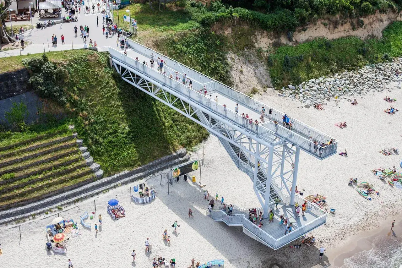 Nowa kładka na plaży Trzęsacz ułatwia dostęp do morza. Ludzie spacerują po niej, podziwiając widoki.