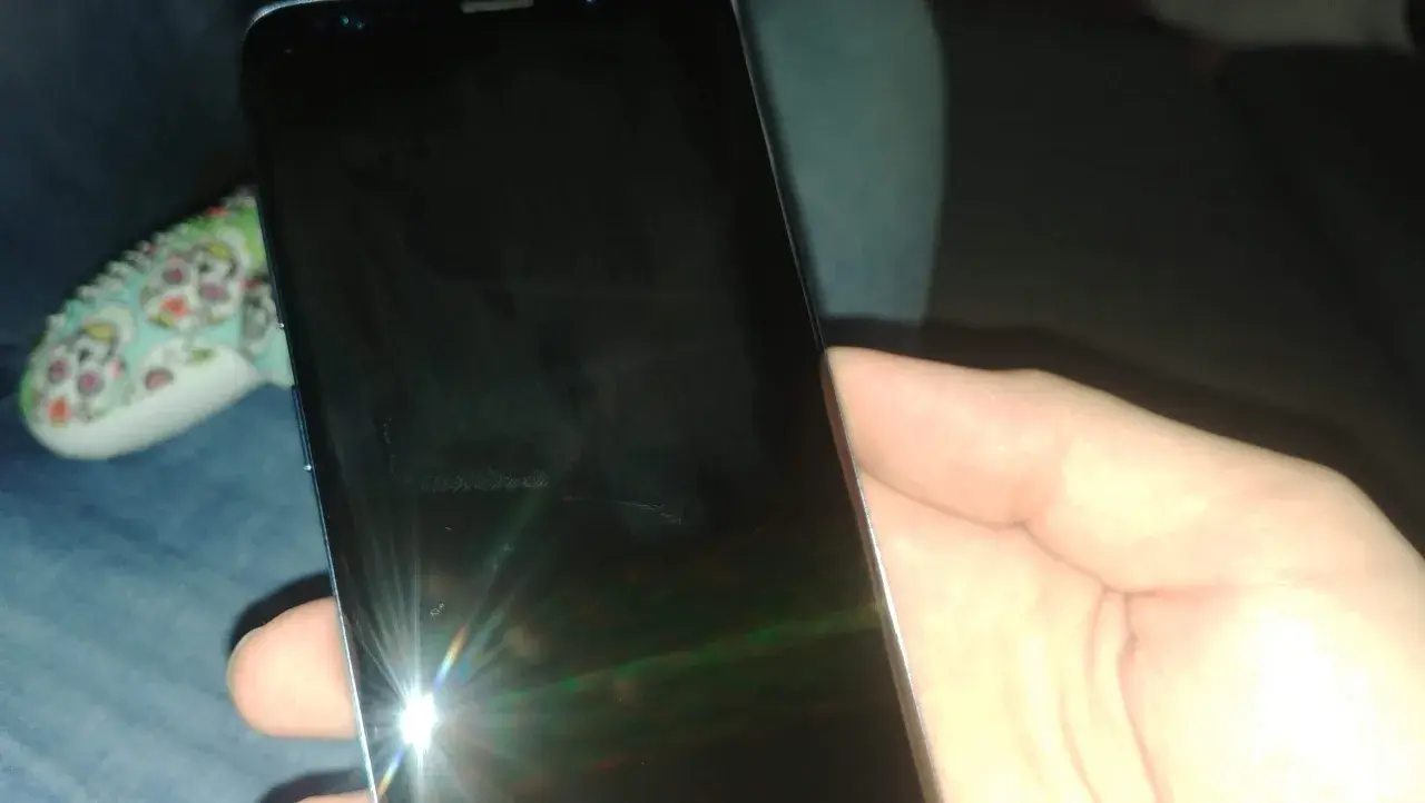 Dłoń trzyma czarny telefon, na którego ekranie widać odbicie światła i ledwo widoczną kreskę na telefonie.