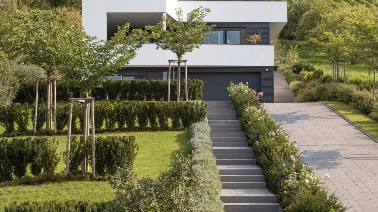 Moderner Garten am Hang mit gepflasterter Auffahrt, Treppen und gepflegten Hecken. Ein Haus mit weißen Fassaden und dunklen Fenstern dominiert den Hintergrund.