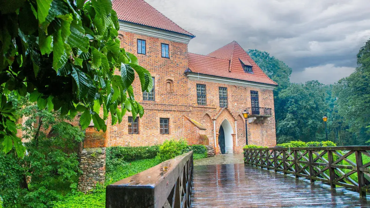 Deszczowy dzień nad zamkiem w Oporowie. Drewniany mostek prowadzi do ceglanego budynku, otoczonego zielenią.