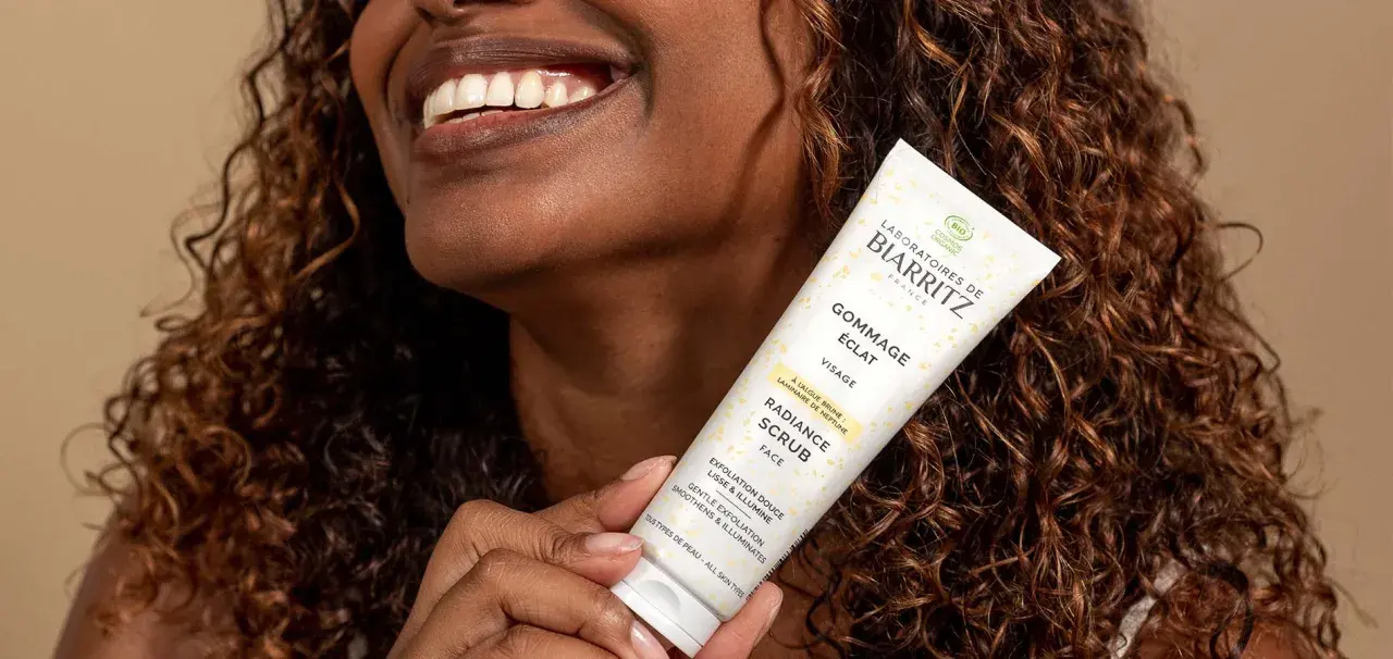Une femme souriante tient un tube de gommage visage éclat. Ce produit sert à lisser et illuminer la peau, révélant sa beauté naturelle.