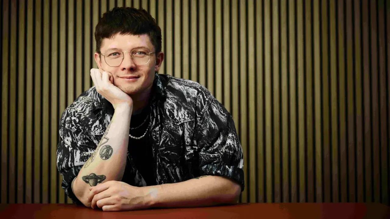 Fritz Espenlaub, mit Brille und Tattoos, lehnt nachdenklich am Tisch vor einer vertikalen Wand.