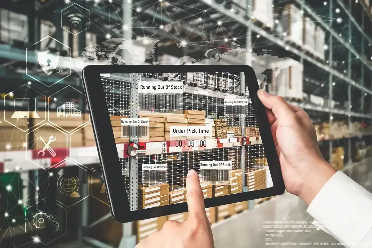Ręce trzymają tablet z widokiem magazynu, pokazujący alerty "Running Out Of Stock" i czas kompletacji zamówienia. To przykład nowoczesnego zarządzania łańcuchem dostaw.
