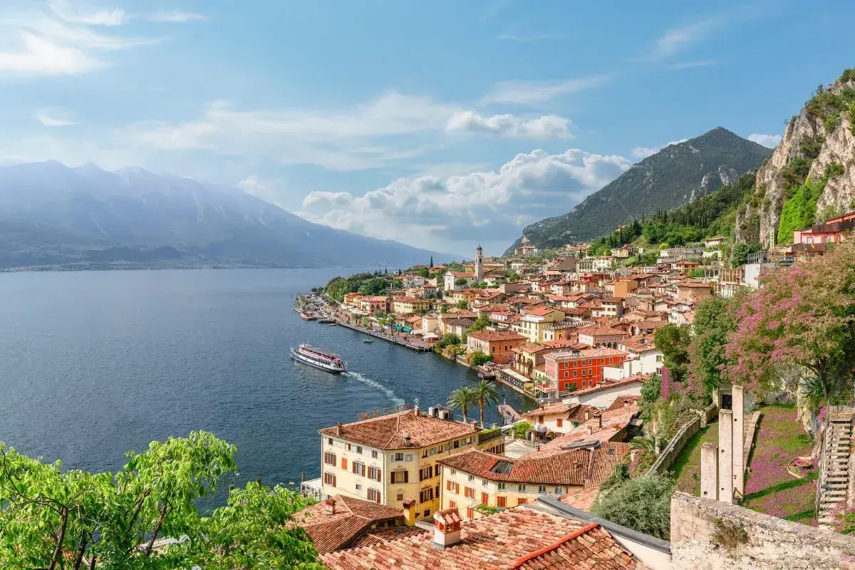 Vista panoramica di una città con tetti rossi e case colorate, incastonata tra la montagna e il blu del lago di Garda. Una nave solca le acque tranquille.