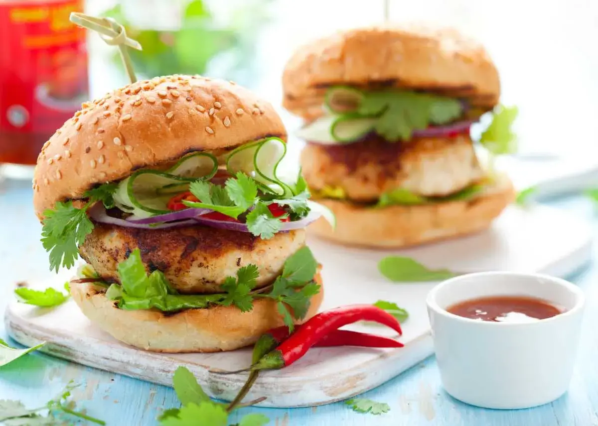Dos hamburguesas de pollo frescas y jugosas, adornadas con pepino, cebolla morada, cilantro y chile rojo.