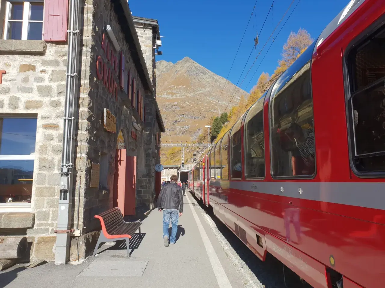 En man går längs en tågstation i bergen, redo för sin tågluffa Europa rutt. En röd tågvagn står parkerad.