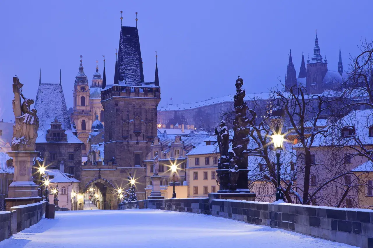 Praga zimą. Śnieg pokrywa Most Karola, wieże i dachy. W oddali widać zarys zamku.