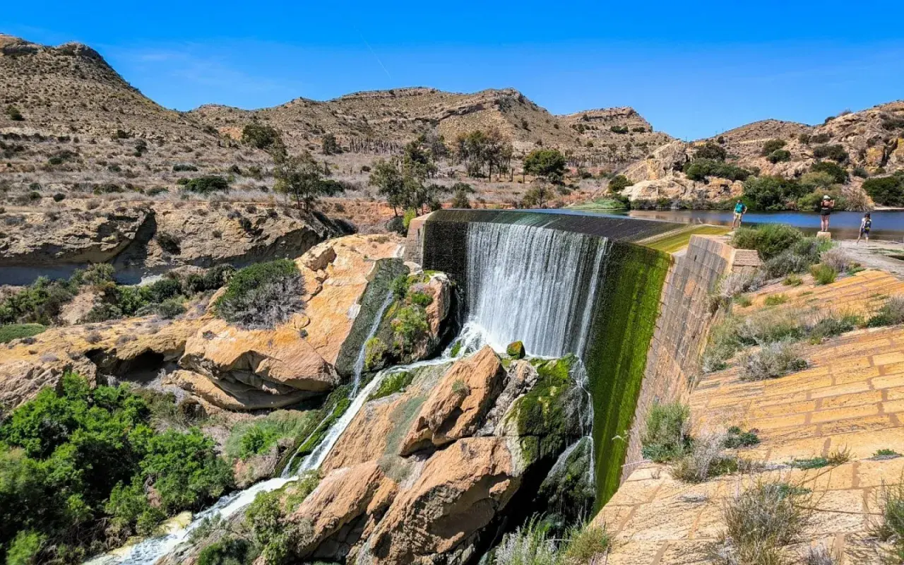 Cascada en el pantano de Elche, con rocas cubiertas de musgo y vegetación verde. Personas observan el agua.