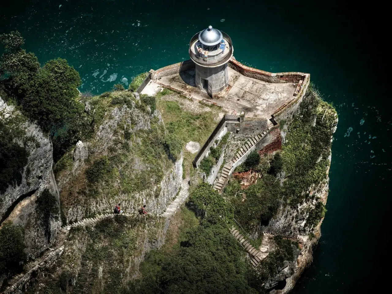Dos personas caminan por un sendero empinado hacia el faro de caballo, un faro solitario en un acantilado rocoso sobre el mar turquesa.