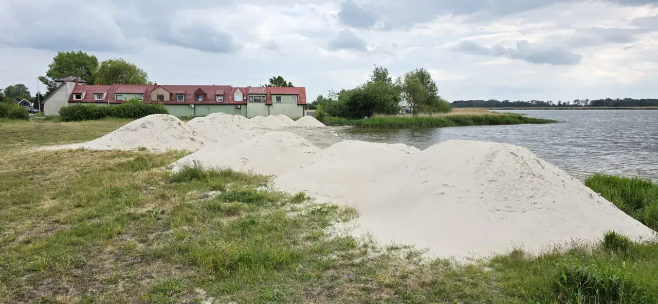 Piasek na plaży w Kamieniu Pomorskim czeka na budowę. W tle zielone budynki i spokojna woda.