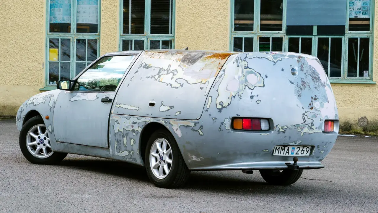 En sliten Saab 600, med rostfläckar och avskavd lack, står parkerad framför en gul byggnad med gröna fönster.