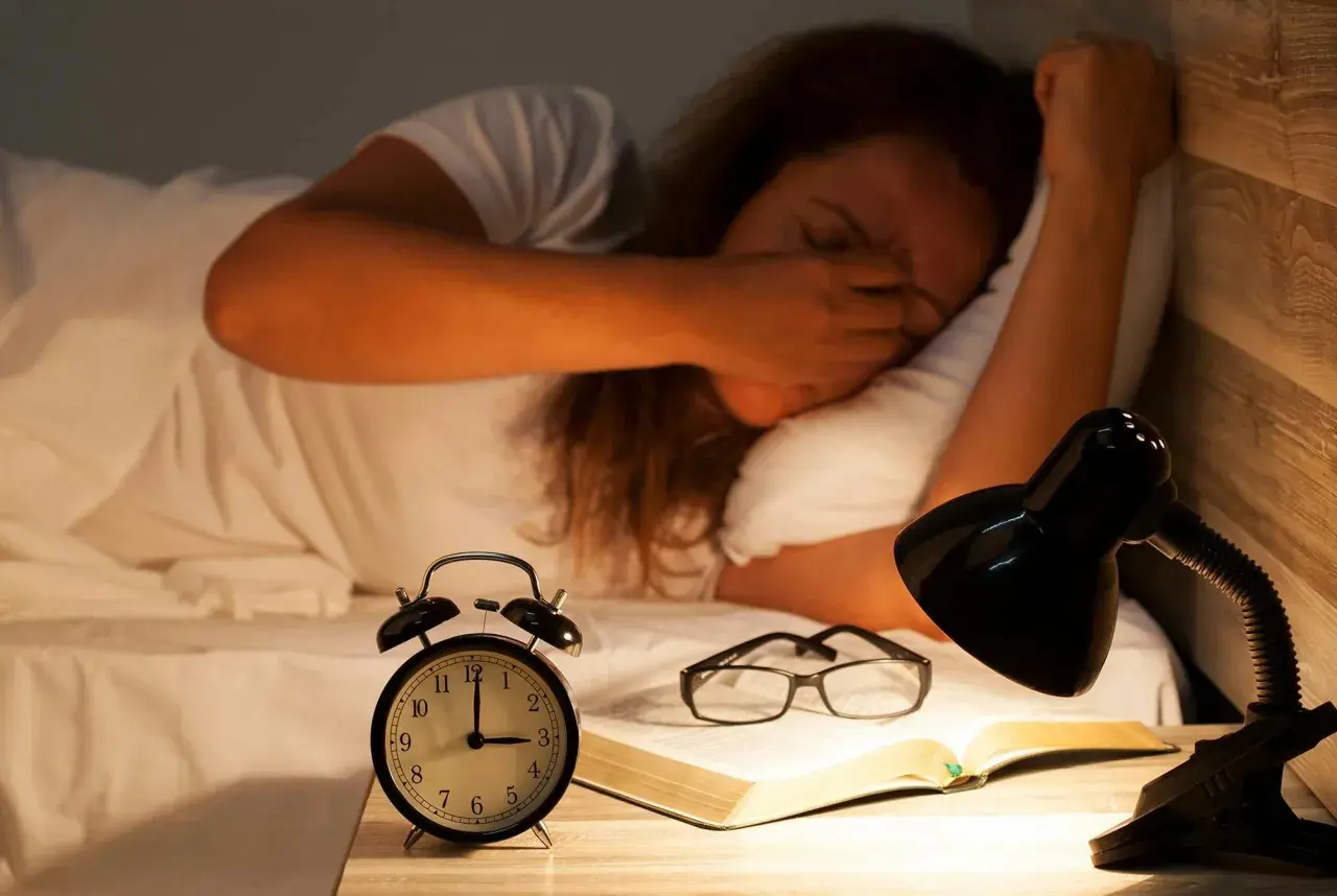 En person som gnuggar sig i ögonen i sängen, med en väckarklocka, glasögon och en bok på nattduksbordet. Känns som att vaknar flera gånger per natt.