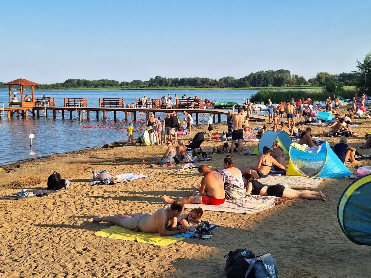Plaża nad jeziorem powidzkim tętni życiem. Ludzie odpoczywają na piasku, kąpią się i spacerują po molo.