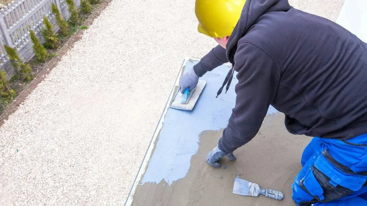 Pracownik w żółtym kasku wykonuje hydroizolację tarasu, nakładając masę izolacyjną na betonową płytę.