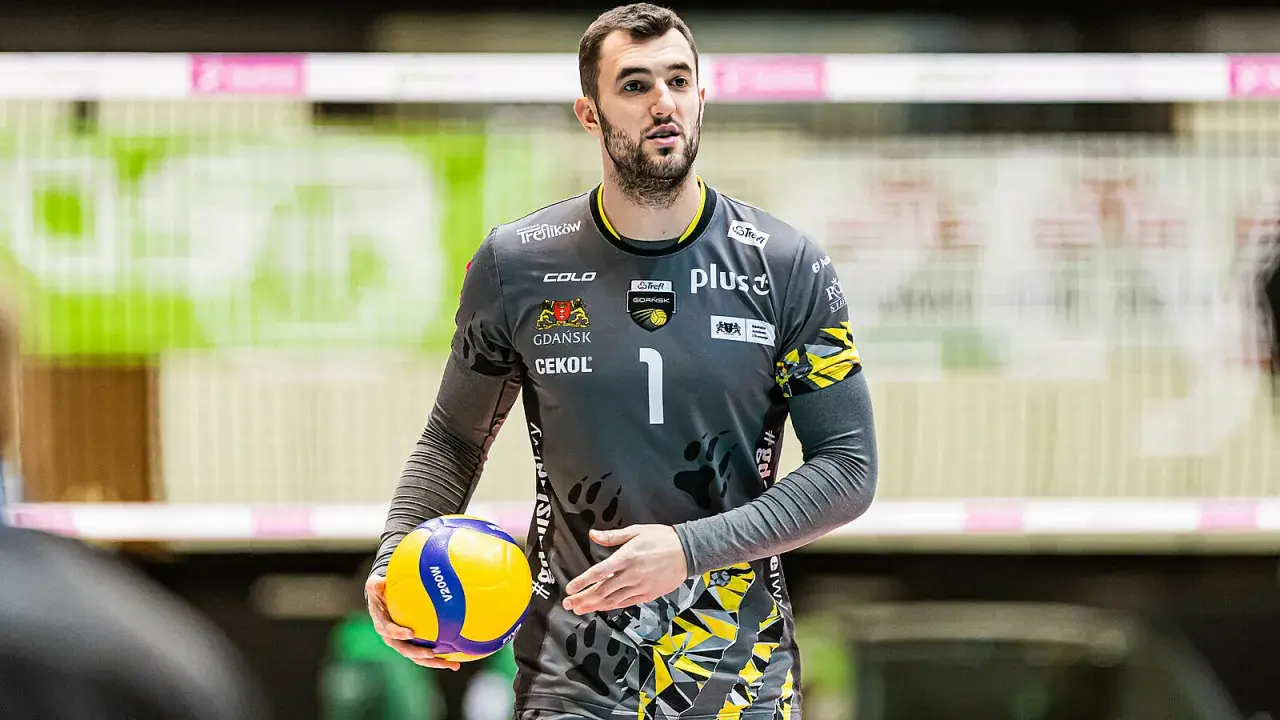 Bartłomiej Lipiński, siatkarz w szarym stroju z numerem 1, trzyma piłkę do siatkówki.