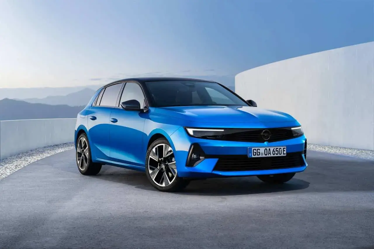 Błękitny opel elektryczny z czarnym dachem, zaparkowany na betonowej nawierzchni, z górzystym krajobrazem w tle.