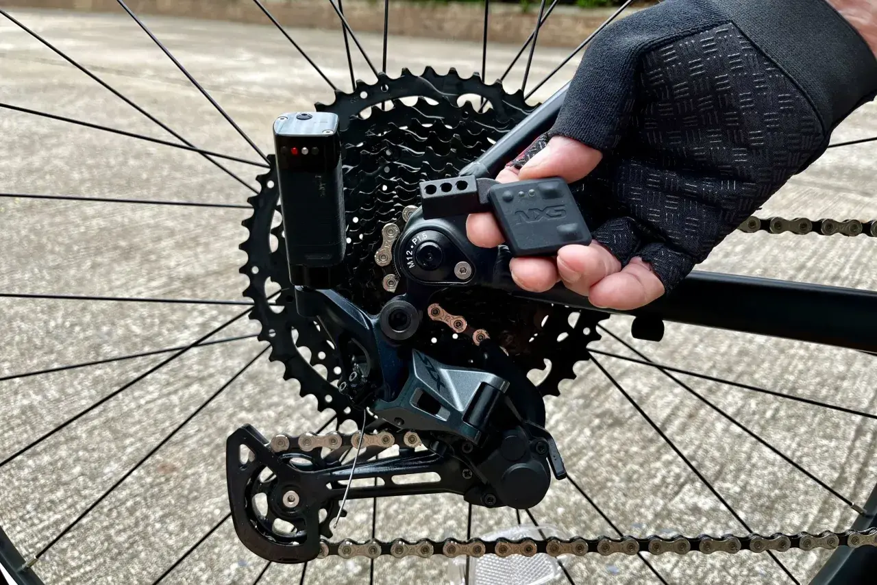 Close-up of a mountain bike's rear derailleur, showing the NX5 electronic shifting system being adjusted by a gloved hand.