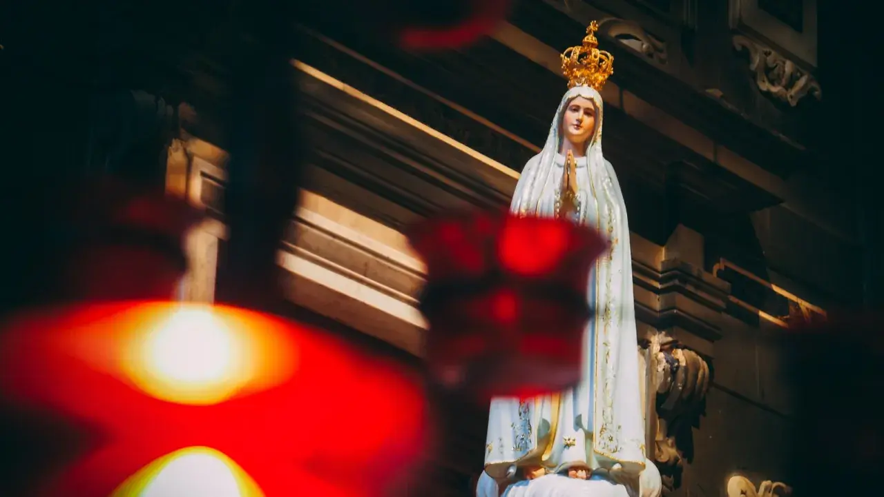 Figurka Matki Bożej Fatimskiej w koronie, z rękami złożonymi do modlitwy. W tle widać architekturę kościoła, a na pierwszym planie rozmyte czerwone światła, symbolizujące trzecia tajemnica fatimska.